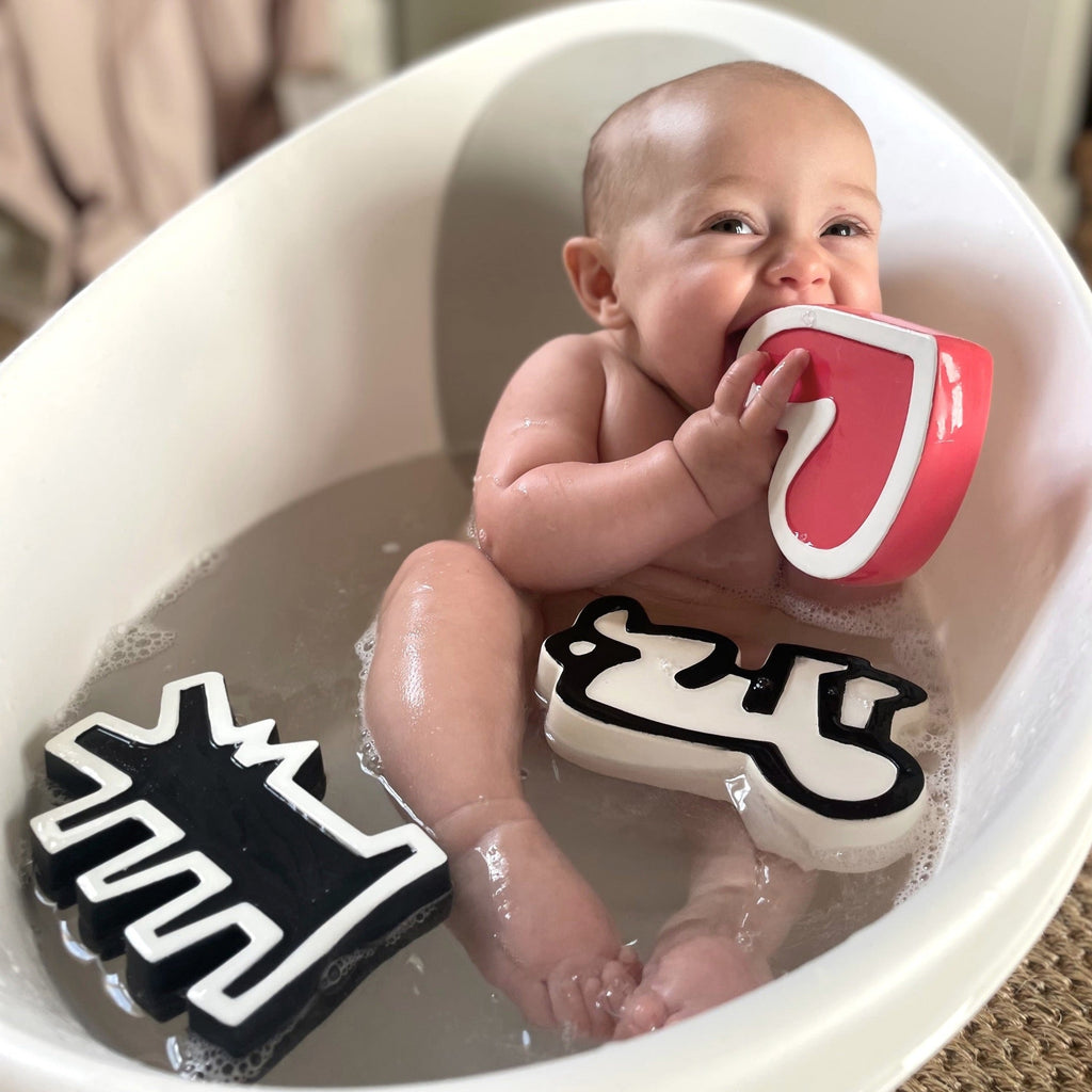 cute baby in a bath tub chewing Keith Haring natural rubber toys - a heart, baby and dog shapes